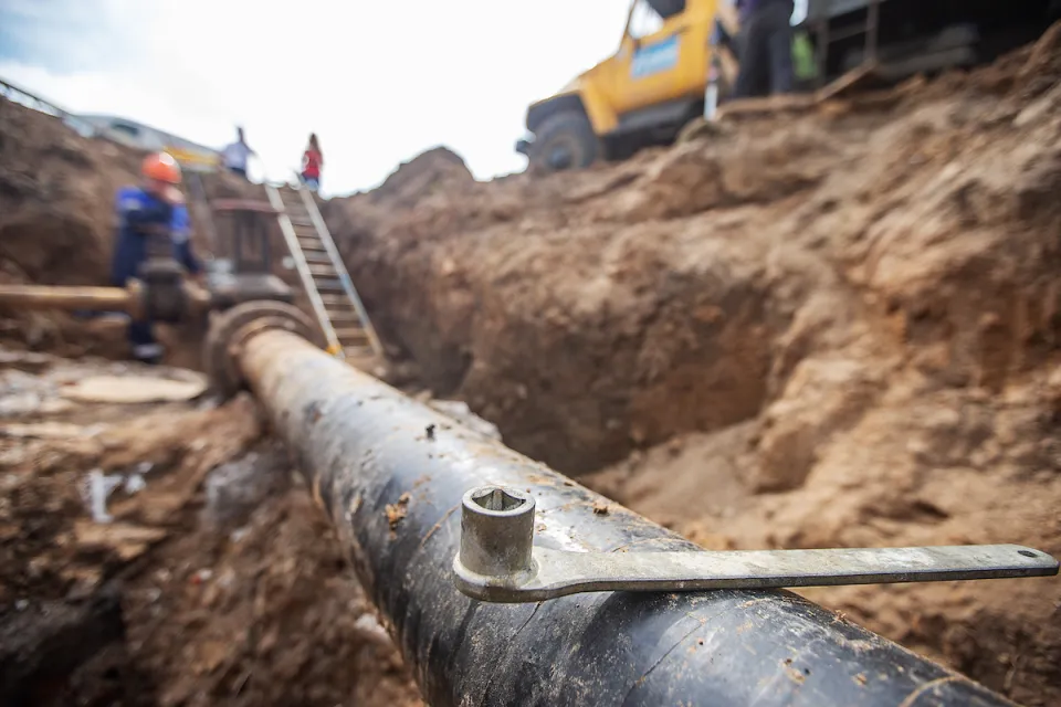 Close-up of exposed pipe in trench with blurred utility workers and yellow truck in background.